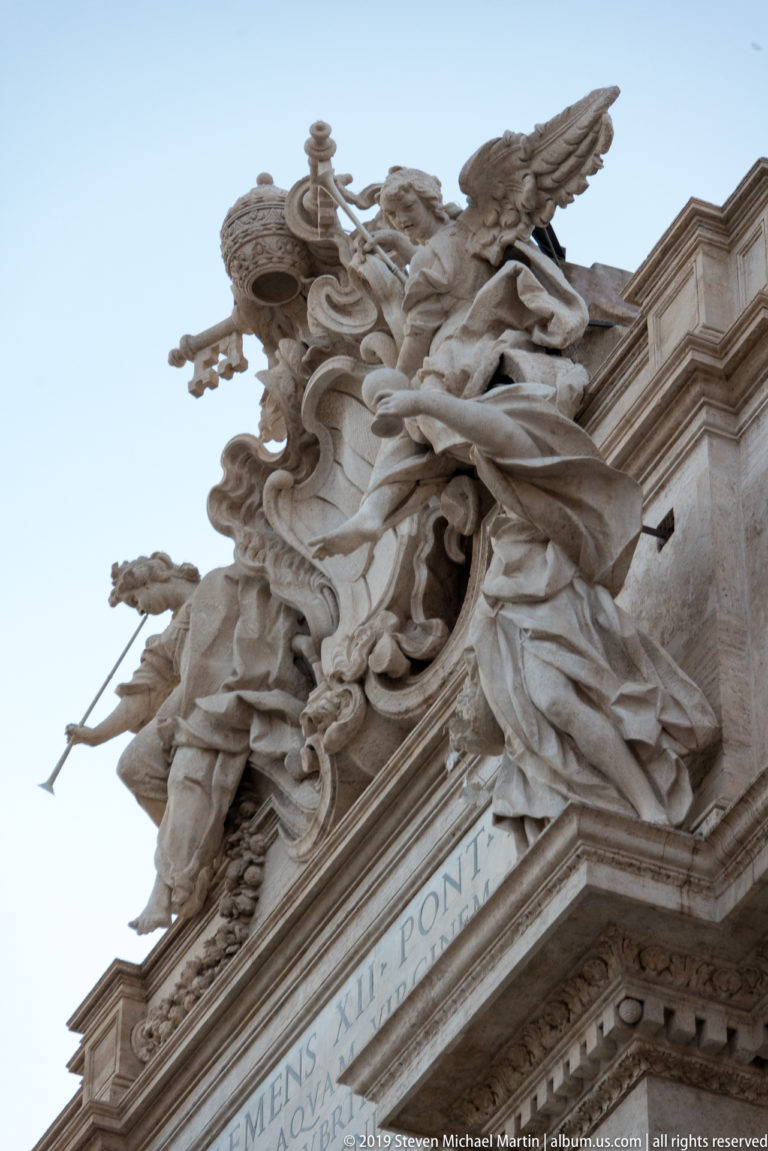 Low-angle detail of Trevi Fountain roofline with papal heraldry, winged figures, scrollwork, and pale stone façade.