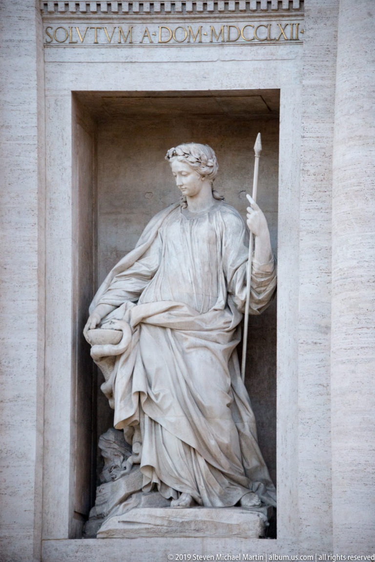 Standing robed female allegorical statue holding a long attribute in a tall niche of the Trevi Fountain.