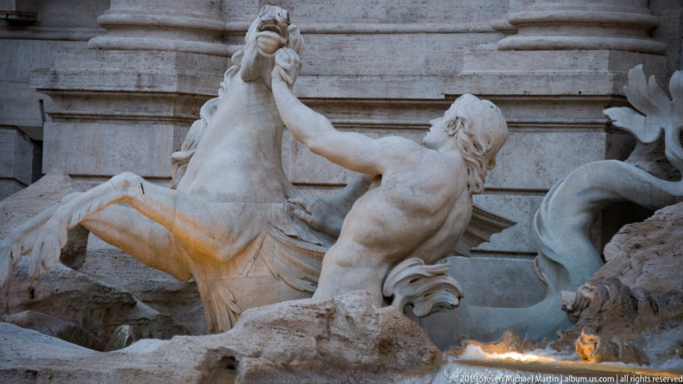 Rearing stone sea horse beside a muscular attendant amid rocky sculpture at the Trevi Fountain.
