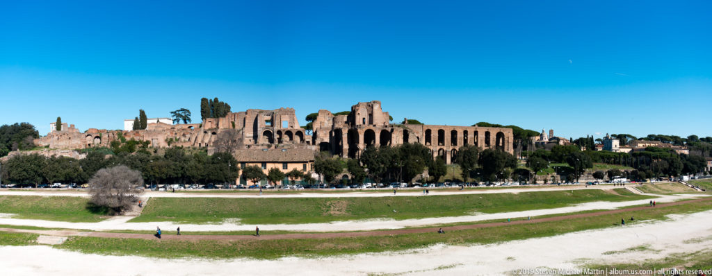 Wide Circus Maximus valley with paths and visitors below extensive brick palace ruins on the Palatine Hill.