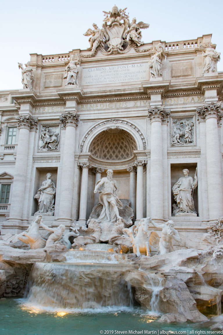Wide view of the Trevi Fountain and Palazzo Poli façade with central niche, statues, columns, and rocky basin.