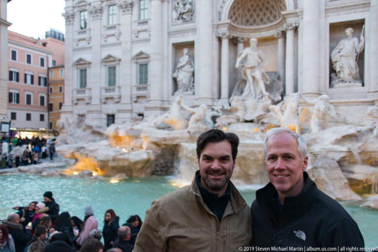 Two men posing in front of the crowded Trevi Fountain, with turquoise water and illuminated Baroque sculpture behind them.