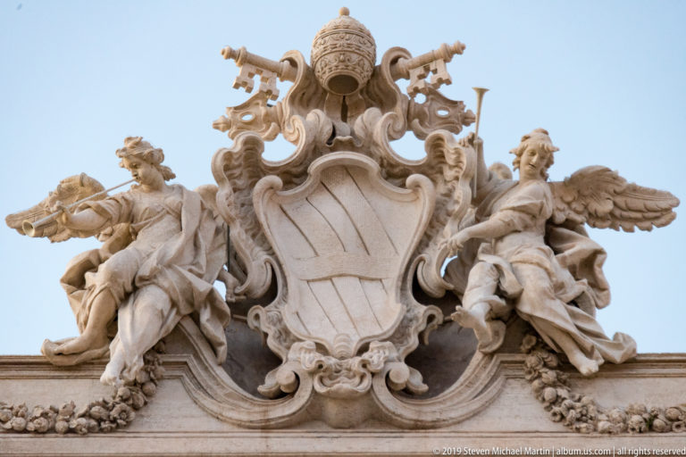 Baroque papal coat of arms with tiara and crossed keys, flanked by two winged figures against the sky.