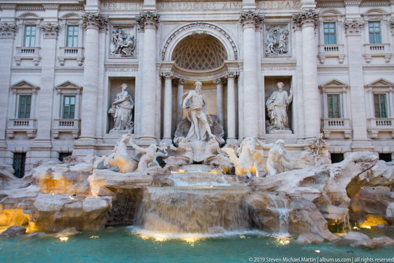 Trevi Fountain at dusk with illuminated sculptures, rocky cascades, pale Baroque façade, and glowing water basin.