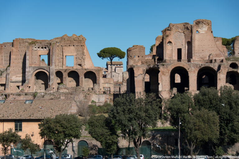 Large brick palace ruins with tall arches and umbrella pines rising on the Palatine Hill above the Circus Maximus.