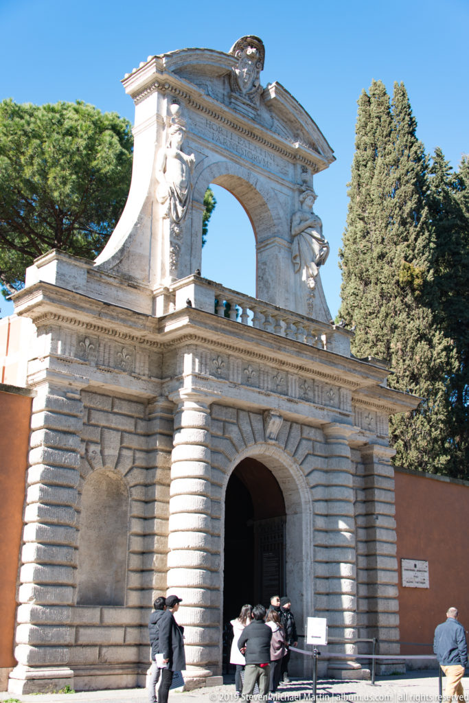 Rusticated stone gate with central passage, large open upper arch, sculptural ornament, and evergreen trees behind.