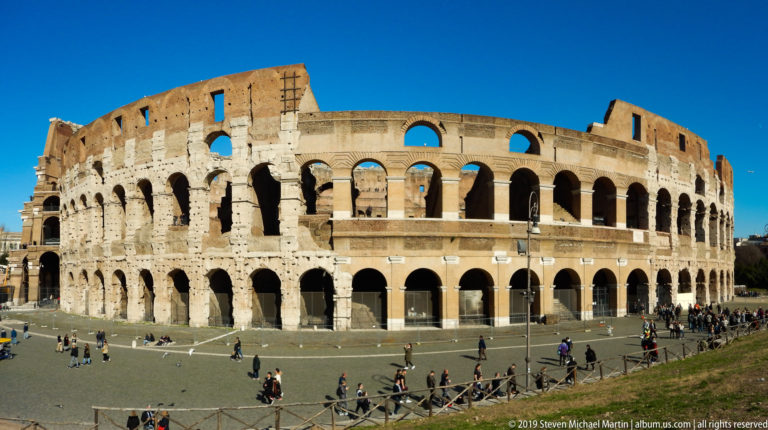 Curved Colosseum façade with multiple tiers of arches, damaged upper wall, and visitors crossing the plaza.