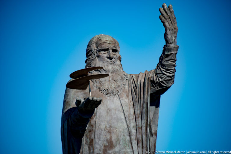 Bronze Leonardo da Vinci holding a model flying machine and raising one hand against a clear blue sky.