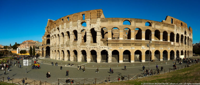 Wide Colosseum exterior with stacked arcades, weathered upper masonry, plaza, and scattered visitors.