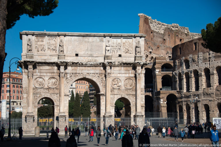 Arch of Constantine centered before the Colosseum's arcades with visitors gathered along the foreground fence.