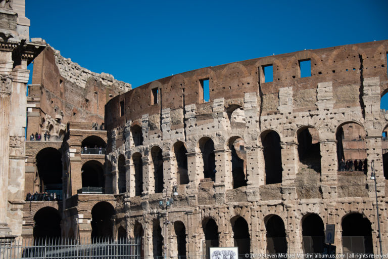 Close exterior of the Colosseum with weathered arcades and broken upper masonry beside the edge of the Arch of Constantine.