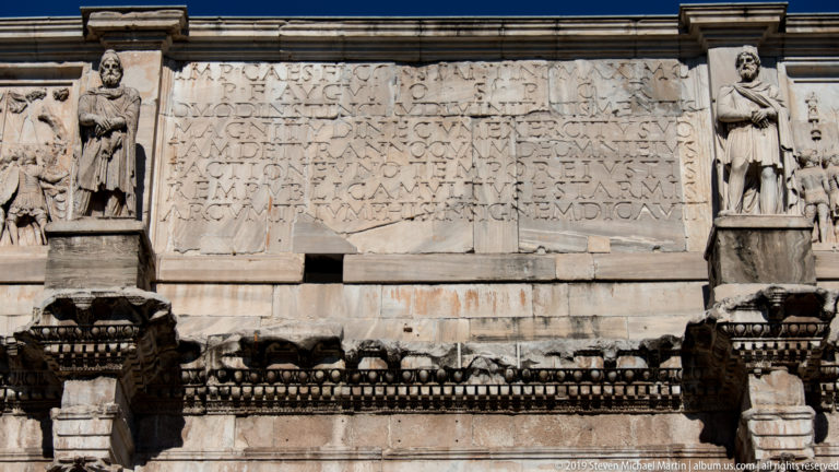 Large Latin inscription on the Arch of Constantine flanked by standing captive statues above projecting architectural moldings.