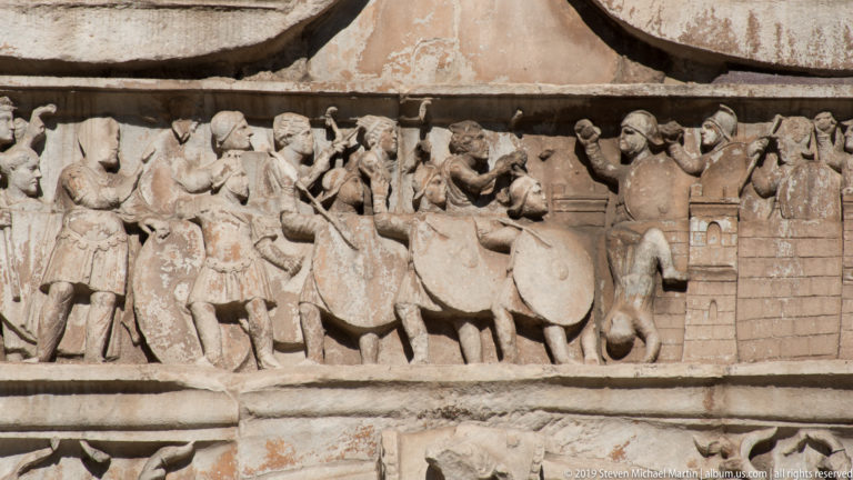 Crowded rectangular relief with soldiers, horses, shields, standards, and architecture on the Arch of Constantine.