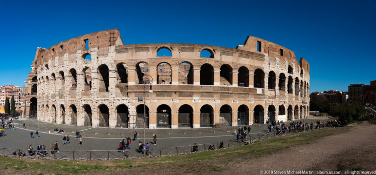 Elevated view of the Colosseum's curved exterior with stacked arches and visitors crossing the surrounding plaza.