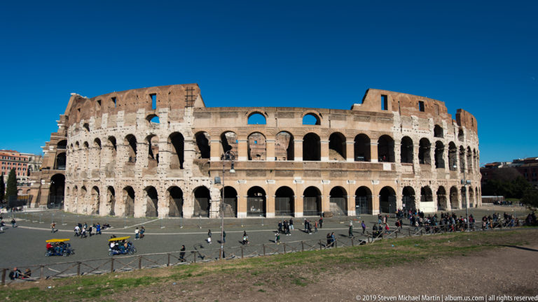 Wide exterior view of the Colosseum with stacked arcades, weathered upper walls, and visitors around the plaza.