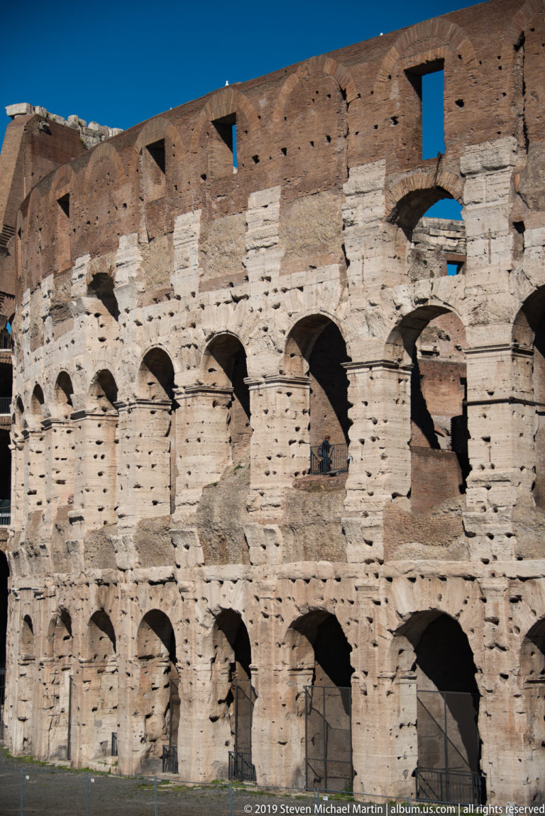 Weathered Colosseum wall with brick infill, travertine blocks, arched openings, and many small holes in the masonry.