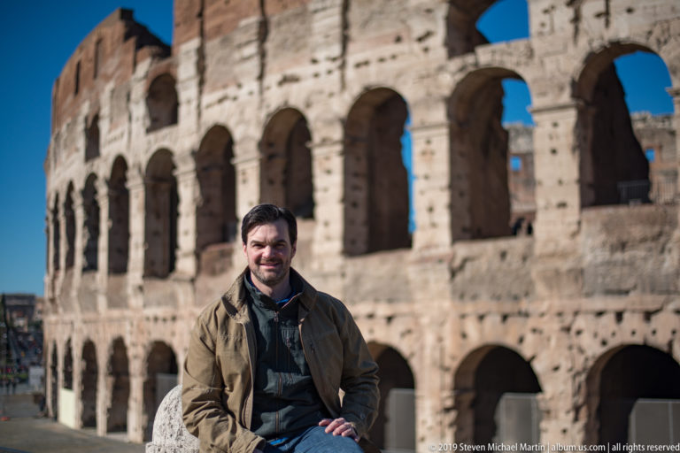 Male visitor seated before several tiers of the Colosseum's curved exterior arcades in bright sunlight.