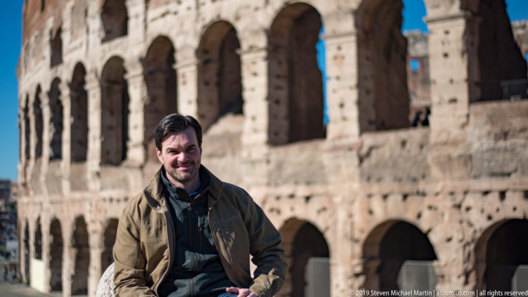 Seated male visitor in brown jacket with the sunlit arches of the Colosseum blurred behind him.