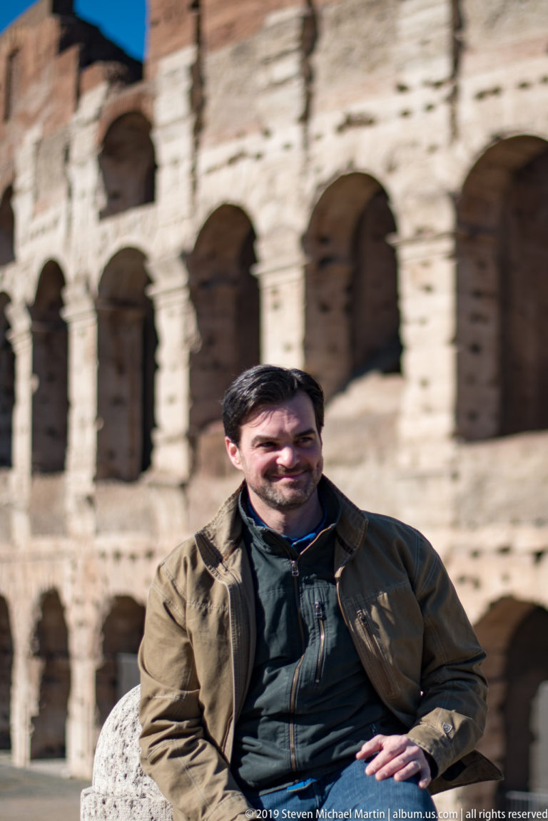 Seated male visitor in a brown jacket with the blurred sunlit arcades of the Colosseum behind him.