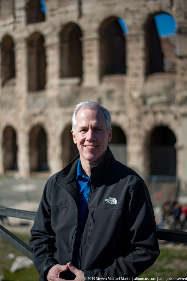 Close portrait of a smiling male visitor with the blurred arches of the Colosseum behind him.