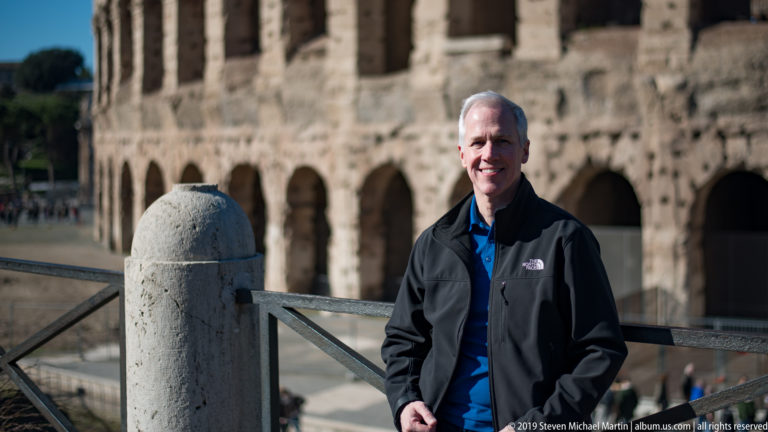Smiling male visitor in dark jacket posing beside a railing with the Colosseum arcades behind him.