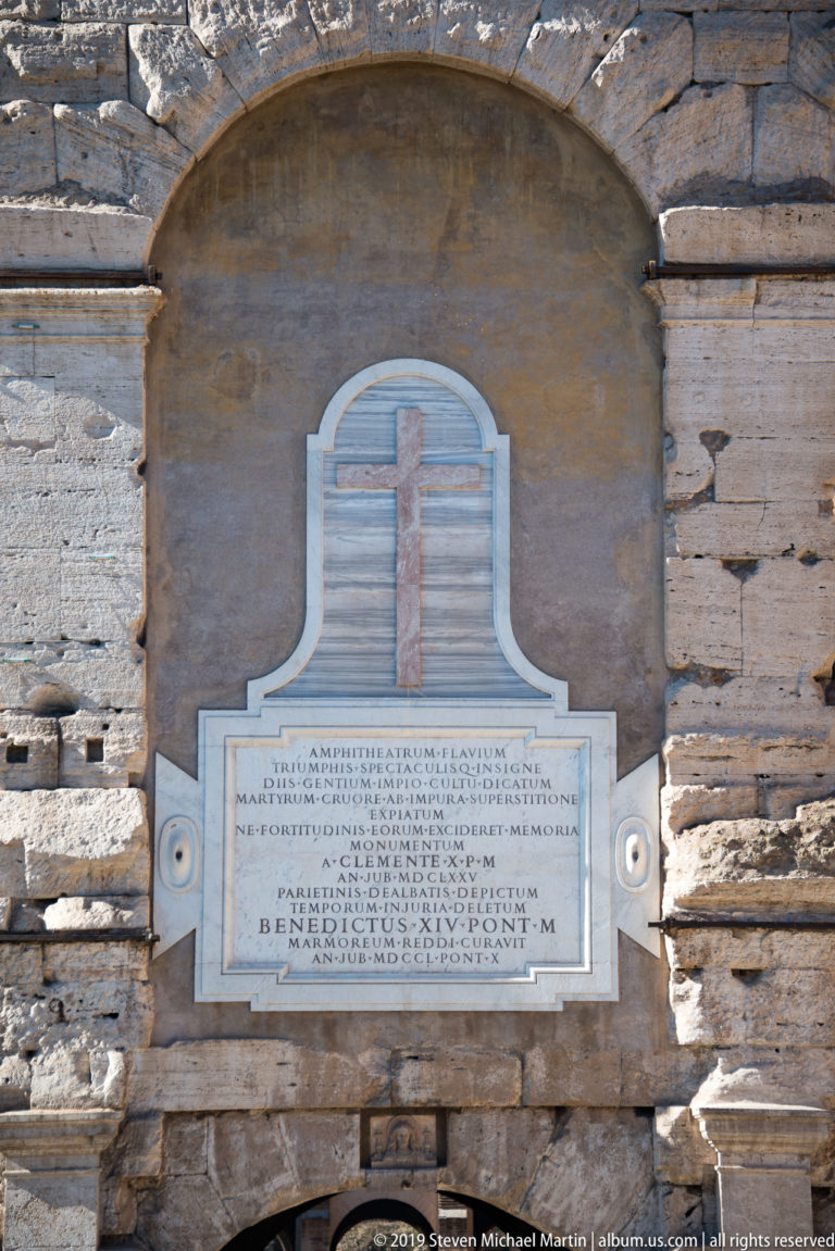 Marble Latin commemorative plaque with a cross mounted within a shallow arched recess of weathered masonry.