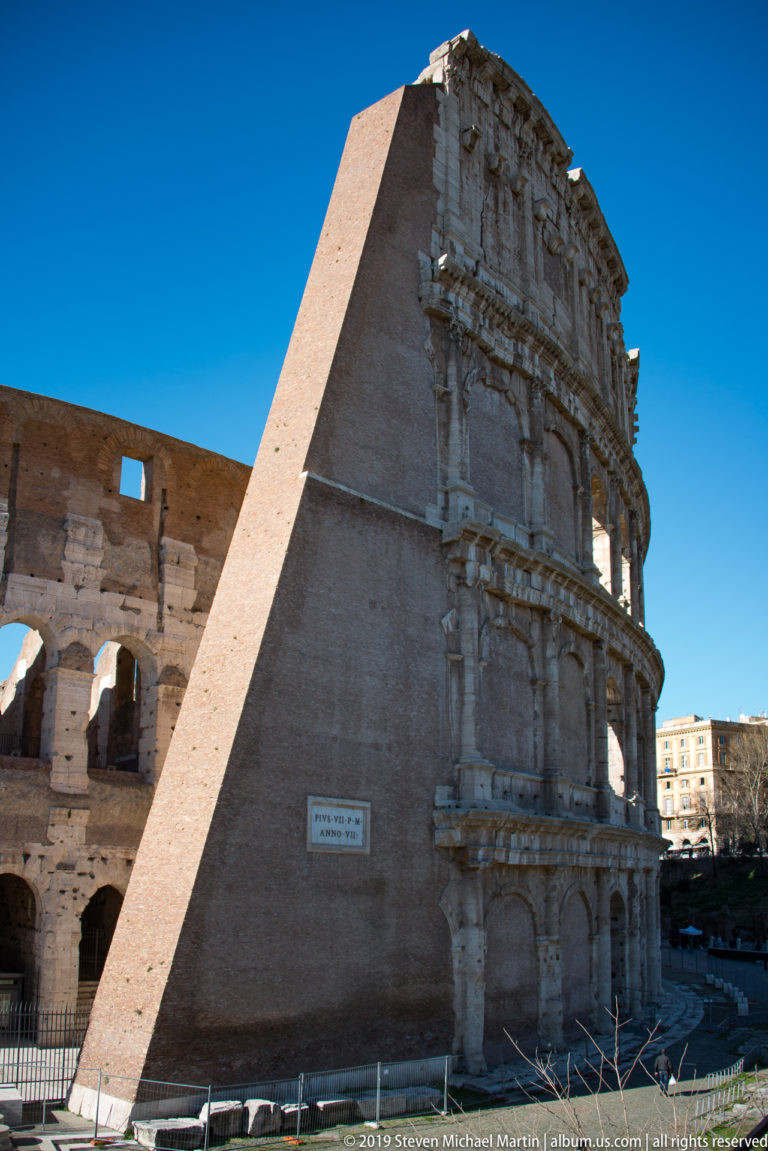 Large brick buttress supporting the exposed end of the Colosseum's weathered travertine arcades under blue sky.