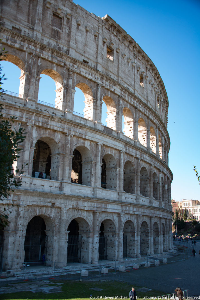 Curved Colosseum exterior with three levels of arches and weathered pale stone under a clear blue sky.