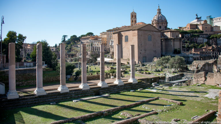 Row of ancient columns above grassy foundations and ruins, with historic churches and Capitoline buildings behind.