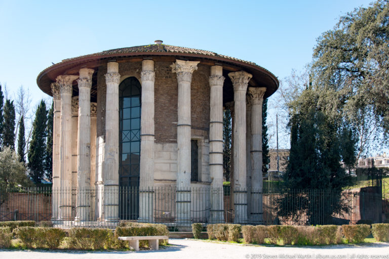 Circular ancient Roman temple with Corinthian columns, tiled roof, fenced garden, and trees under a clear blue sky.