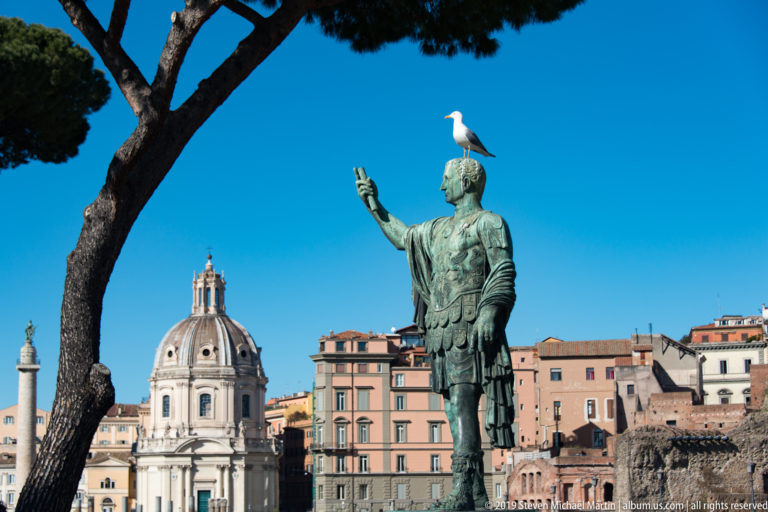 Bronze Augustus raising one hand, gull on his head, with Santa Maria di Loreto and Trajan's Column behind.