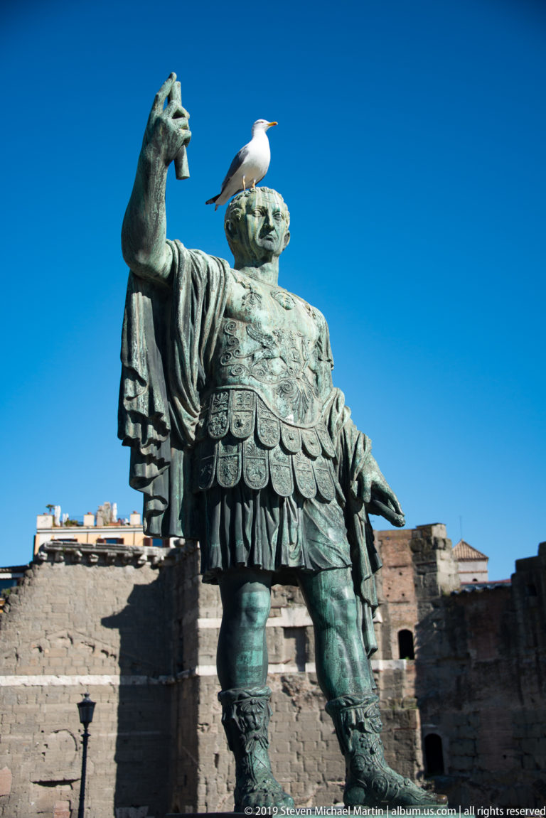 Bronze Augustus in armor raising one hand, with a gull perched on his head before ancient brick ruins.