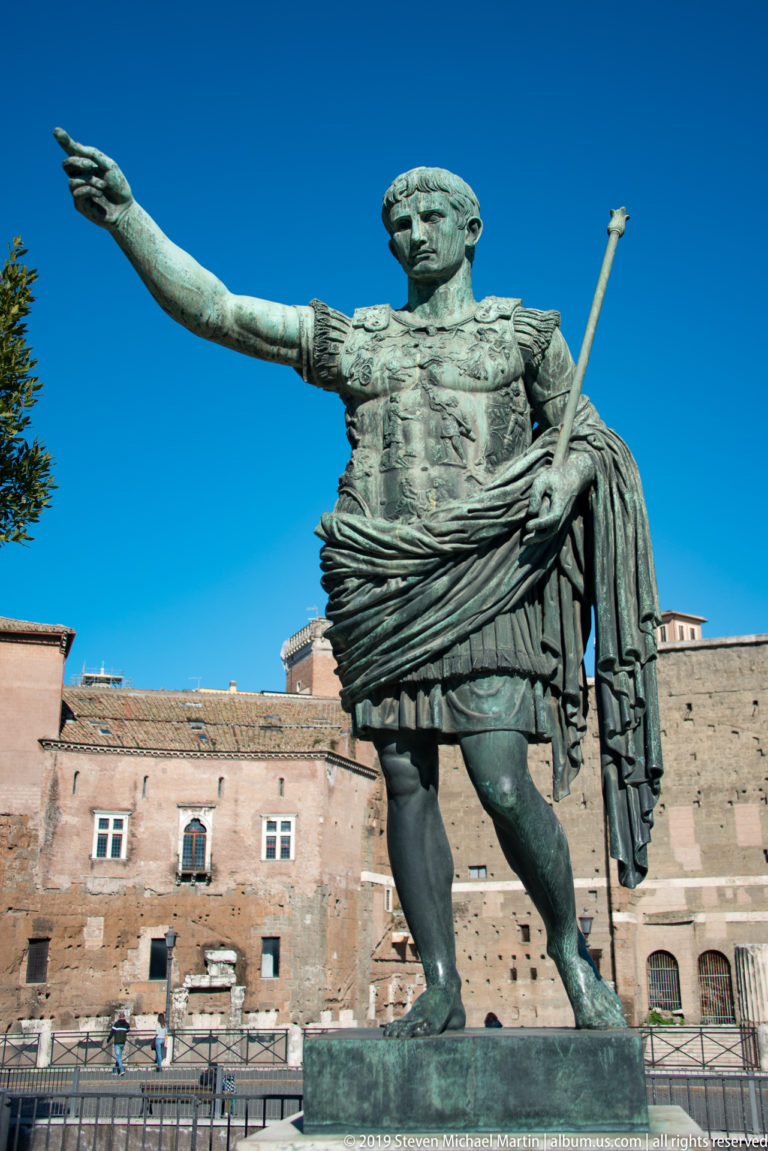 Bronze Emperor Nerva in military dress extending one arm and holding a staff before ancient brick ruins.