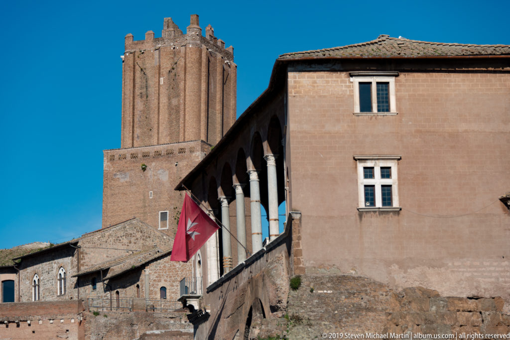 Brick medieval tower behind a warm stucco building with columned loggia and red flag near Rome's imperial forums.