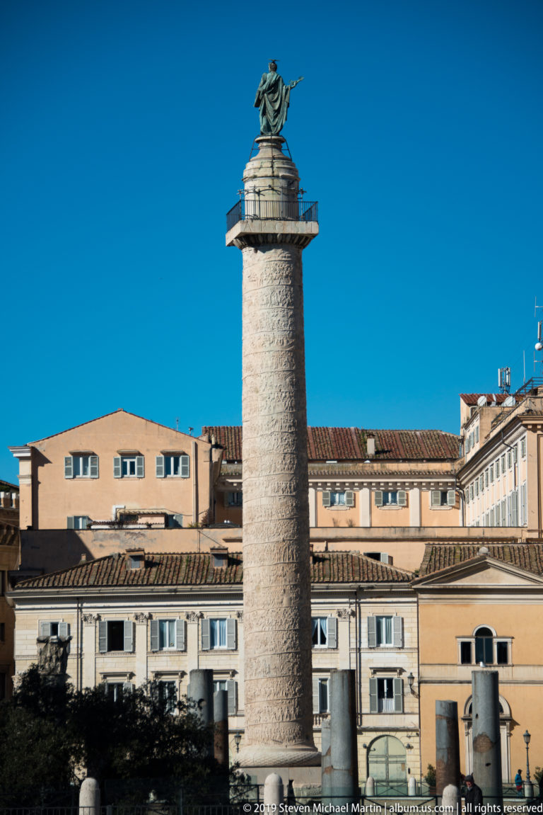 Trajan's Column with spiral relief and Saint Peter statue rising above warm-toned buildings and forum ruins.