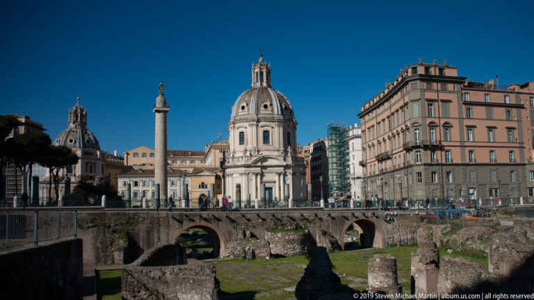 Trajan's Column and domed churches beyond excavated Roman arches, foundations, and masonry in the imperial forums.