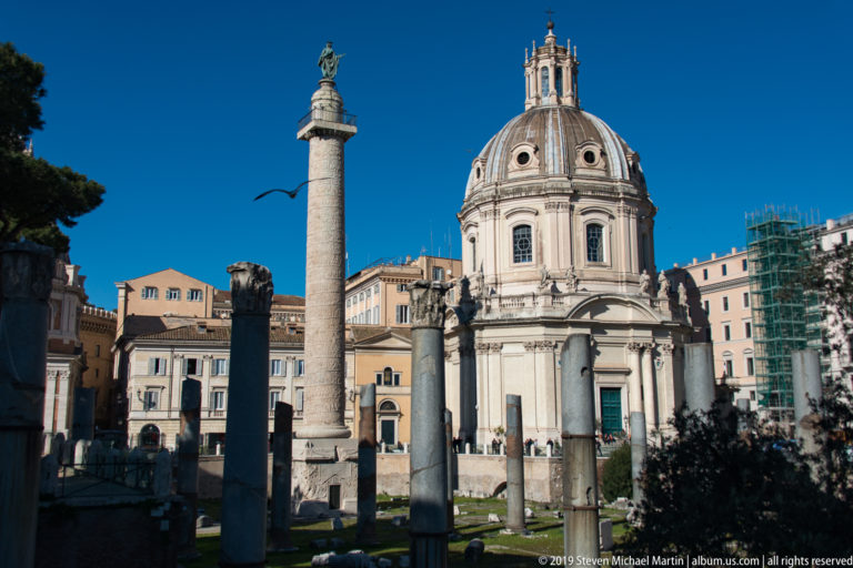 Trajan's Column and domed Santa Maria di Loreto behind ancient columns and archaeological remains in central Rome.