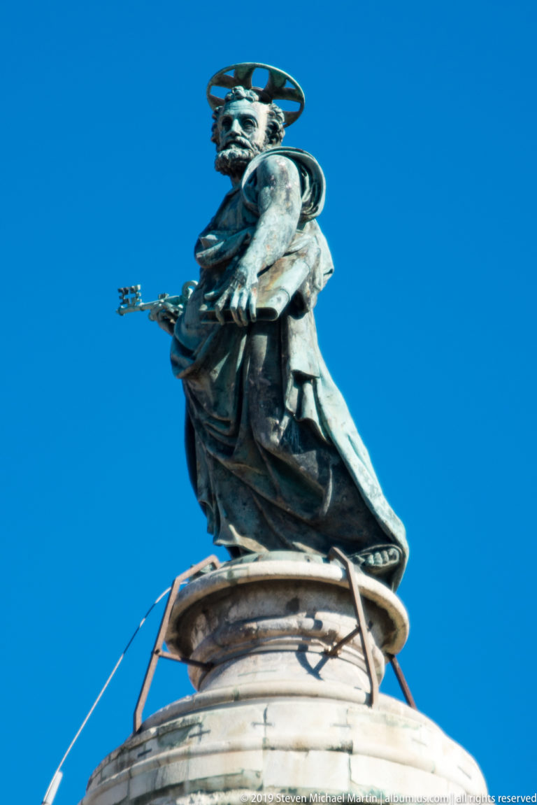 Bronze Saint Peter with halo and keys standing on the summit of Trajan's Column against blue sky.
