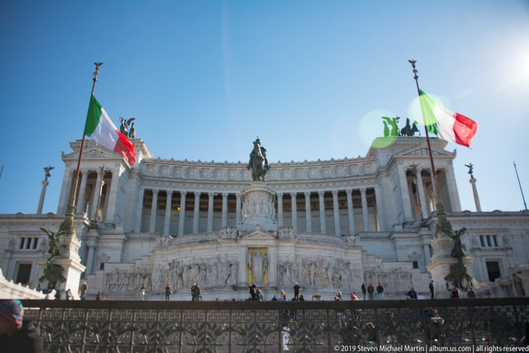 White Vittoriano monument with Corinthian colonnade, equestrian statue, broad stairs, and Italian flags.