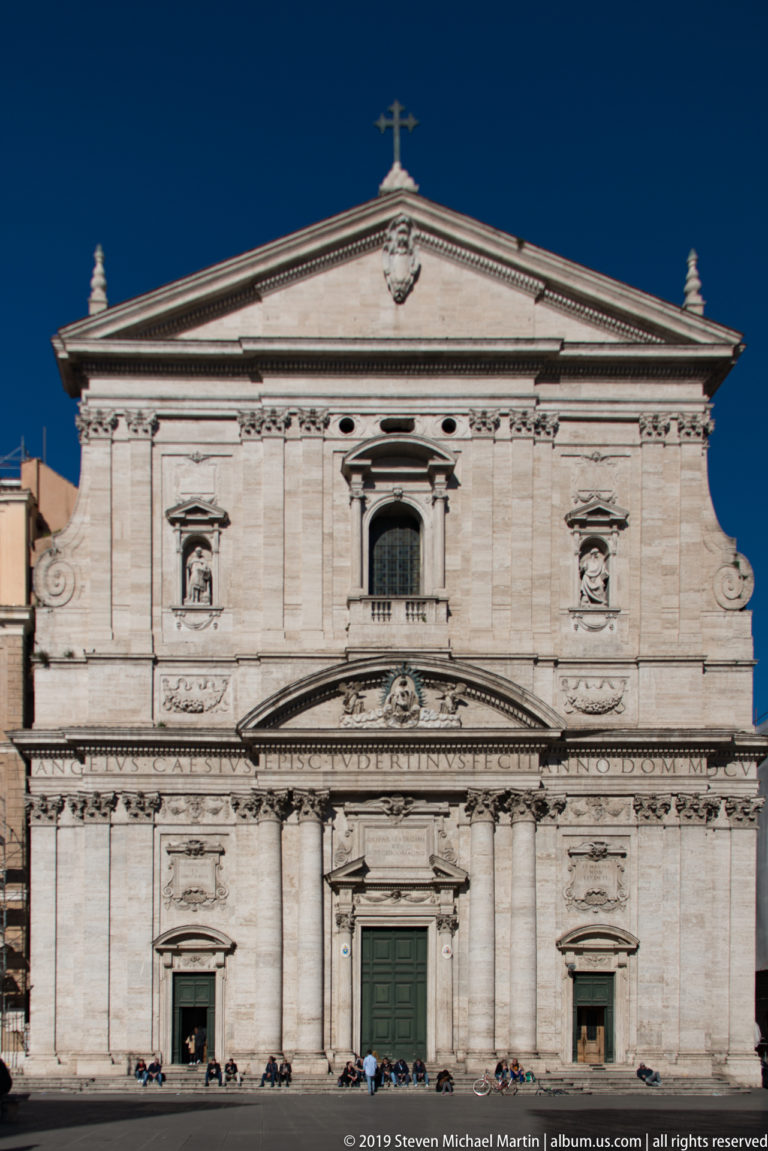 Pale two-story church façade with pilasters, niches, volutes, triangular pediment, and cross under a deep blue sky.