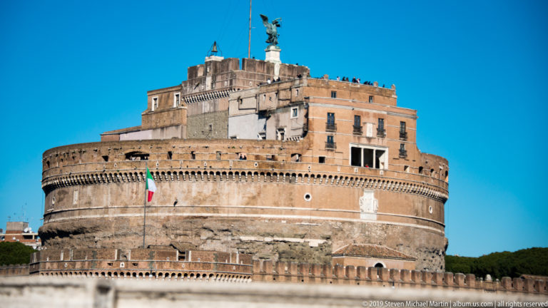 Massive cylindrical Castel Sant’Angelo with defensive walls, Italian flag, upper terraces, and bronze angel on the summit.