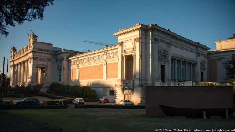 Side view of the National Gallery with warm brick panels, pale classical stonework, cornices, and lawn.