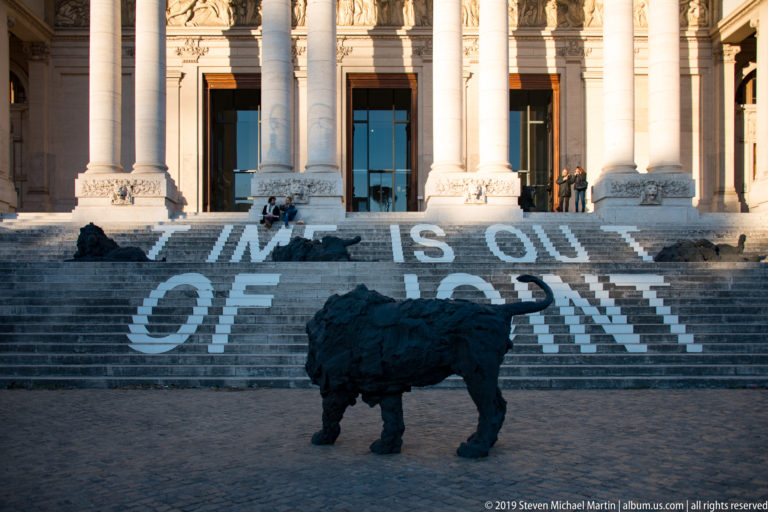 Dark buffalo sculpture standing before museum steps painted with large white lettering and tall classical columns.