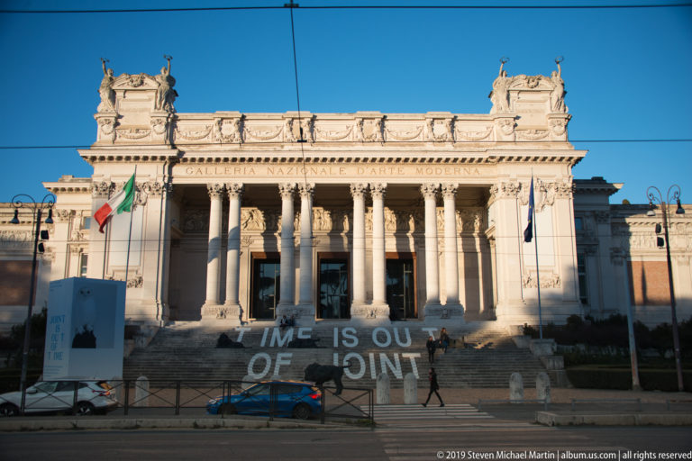 Frontal museum façade with Corinthian columns, Italian flags, broad steps, and contemporary text installation.