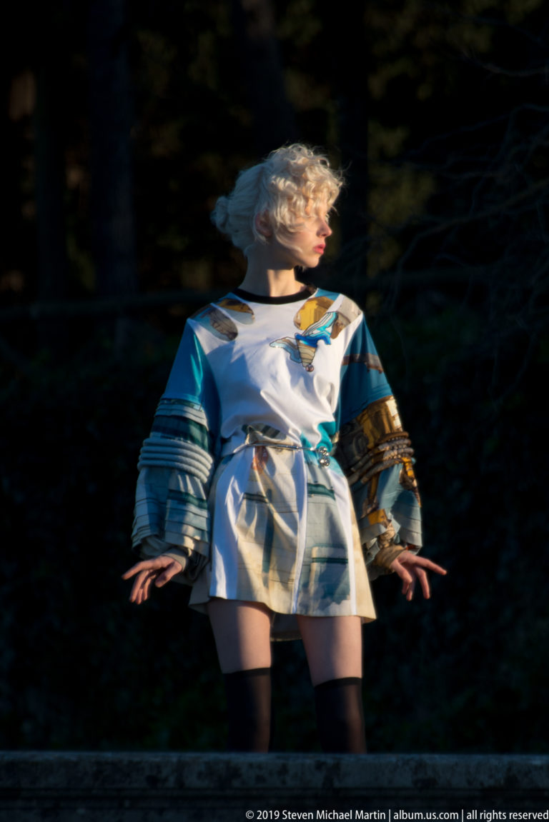 Colorfully patterned female figure sculpture with outstretched arms illuminated against a dark wooded background.