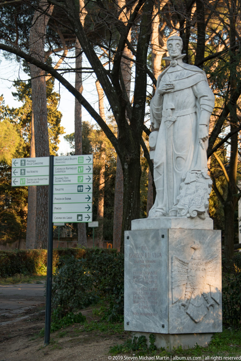 Standing marble figure on an inscribed pedestal beside green directional signs and mature trees in a Roman park.