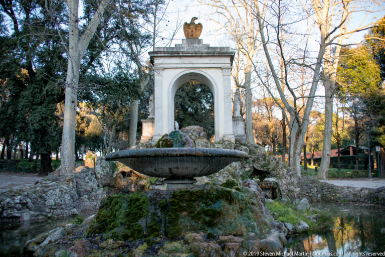 Large stone basin on mossy rocks before a white arched monument crowned by an eagle in a wooded park.