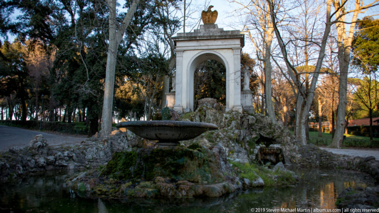Stone basin, mossy rockwork, pond, and white eagle-crowned arch among trees in Villa Borghese.