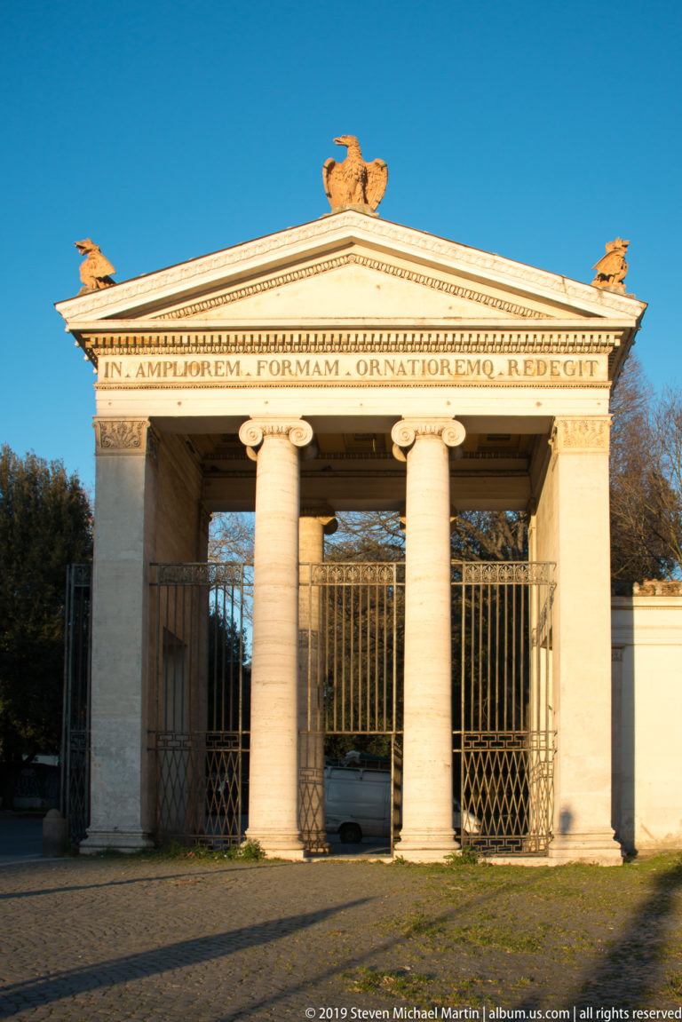 Stone entrance gate with paired columns, triangular pediment, Latin inscription, rooftop sculpture, and closed iron gates.