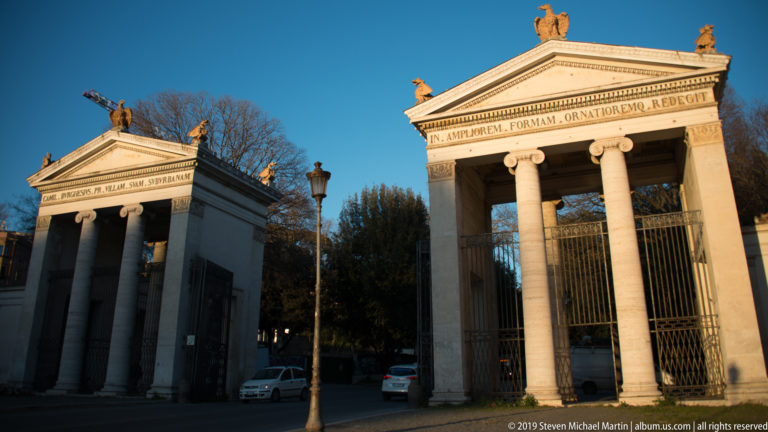 Pair of pale neoclassical gatehouses with columns, pediments, Latin inscriptions, and sculptural roof ornaments.